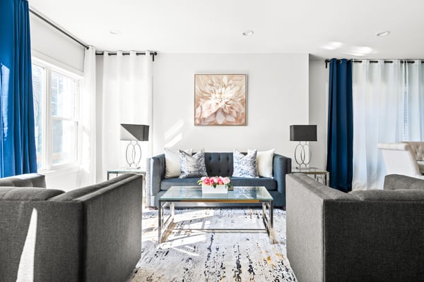 Bright living room with gray armchairs, dark blue sofa, glass coffee table, and blue curtains framing windows