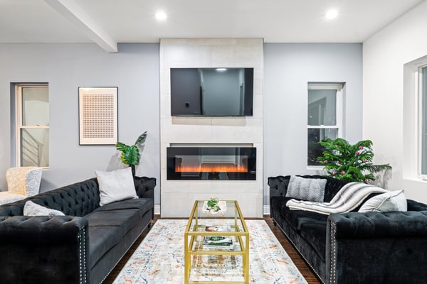 Modern living room with two black tufted sofas, white fireplace with electric insert, wall-mounted TV, gold glass coffee table, and potted plants