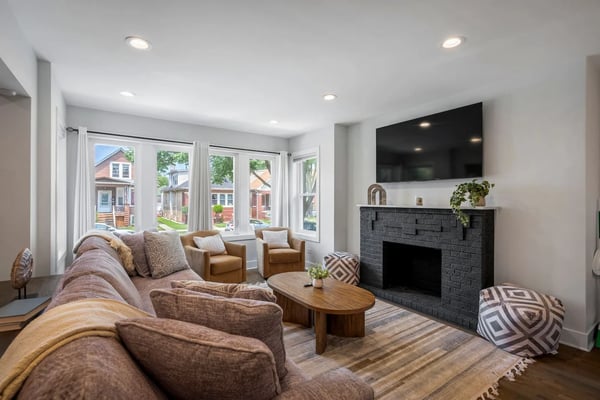 Modern living room with brown sectional sofa, fireplace with black brick surround, wall-mounted TV, and large windows overlooking residential street.