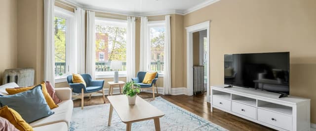 Bright living room with beige walls, bay windows, blue armchairs, white sofa, wooden coffee table, and flat-screen TV on white media console.